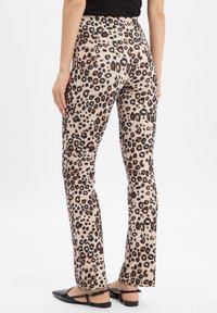 Beige broek met luipaardprint, voorzien van zwarte en bruine vlekken, getailleerd ontwerp, zachte stof, flared pijpopening, gedragen met zwarte platte sandalen.