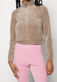 Avklippt beige sammetjacka med hög krage, frontdragkedja och sidofickor. Har en silverlogotyp på bröstet. Matchas med rosa sammetbyxor.