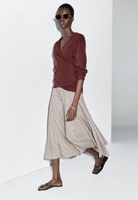 Bordeauxrode wikkeltrui, zachte stof; vloeiende midi-rok met beige strepen; zwarte platte sandalen; gedragen met donkere ronde zonnebril; minimalistisch ontwerp.