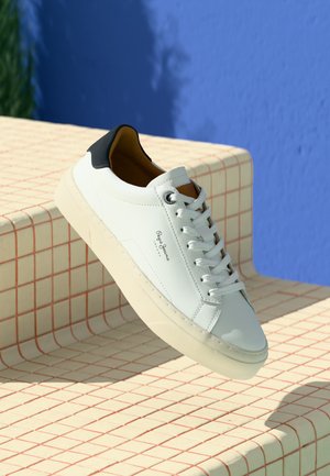 White leather sneaker with black heel tab and white laces floating above cream tiled steps against a blue wall background.