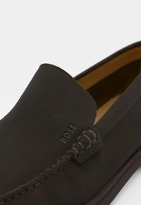 Chaussures en cuir marron à enfiler avec une texture lisse, bout rond et accents cousus. Logo "BOSS" embossé sur le côté.