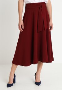Jupe midi bordeaux en tissu doux, avec une taille nouée, un design A-line fluide et une texture lisse. Portée avec des talons navy.