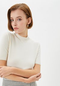 Sweter z krótkim rękawem w kolorze ecru z prążkowanym dekoltem. Modelka ma jasnobrązowe włosy i nosi złote kolczyki w kształcie kół, z założonymi rękami.