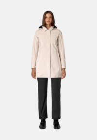 Cappotto impermeabile rosa chiaro con zip frontale, colletto alto e cappuccio blu navy. Indossato sopra pantaloni scuri e scarpe nere.