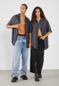 Due persone posano indossando camicie grigio scuro con bottoni. Una abbina la camicia a jeans a gamba larga di colore blu chiaro, l'altra a una gonna nera con spacco alto e stivali.