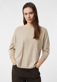 Beżowy sweter z dzianiny z szerokim dekoltem i rękawami do połowy łokcia. Teksturowana tkanina i luźny krój. W połączeniu z ciemnobrązowymi spodniami.