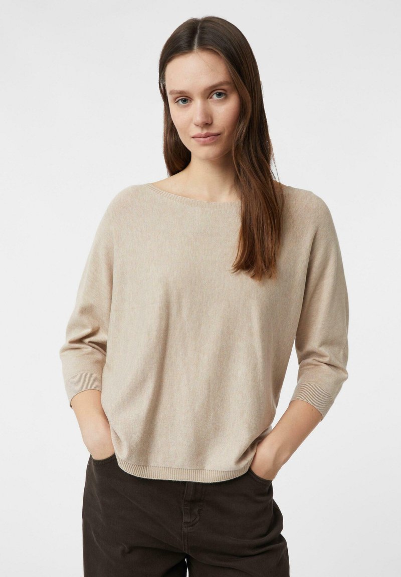 Beżowy sweter z dzianiny z szerokim dekoltem i rękawami do połowy łokcia. Teksturowana tkanina i luźny krój. W połączeniu z ciemnobrązowymi spodniami.