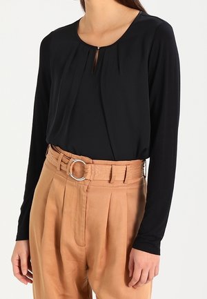 Haut noir à manches longues avec un décolleté arrondi et un détail en forme de clé, associé à un pantalon beige taille haute avec une large ceinture et une boucle en argent.