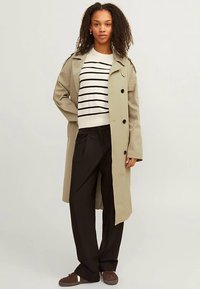 Beżowy płaszcz trencz z podwójnym rzędem guzików, czarne guziki, na białym swetrze w paski, zestawiony z czarnymi spodniami i brązowymi butami.