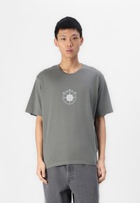T-shirt in cotone grigio con maniche corte, caratterizzato da un design circolare di una bussola bianca al centro. Vestibilità semplice e rilassata.