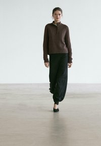 Brown, textured knit pullover with a buttoned collar; paired with loose, black tapered pants and black flats; minimalist fashion look.