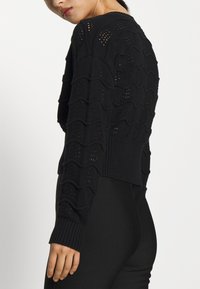 Svart stickad tröja med vågmönster och öppet vävmönster, med ribbade muddar och fåll, mittsnittad längd.