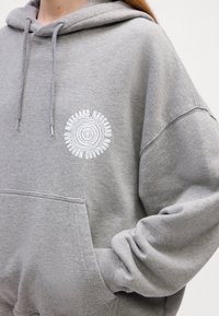 Svetlo šedá oversized mikina s predným vreckom, šnúrkami a bielym kruhovým logom na hrudi. Mäkká textura, hrubá tkanina.