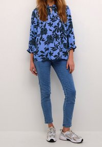 Blouse fleurie bleu clair avec motifs vert foncé, détail avec une ouverture en forme de goutte, et manches à volants, assortie à un jean slim bleu et des baskets grises.