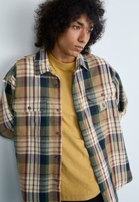 Camisa de franela de gran tamaño en cuadros multicolores (marrón, verde, azul marino) con una camiseta amarilla de cuello redondo debajo, que presenta detalles de botones y un bolsillo en el pecho.