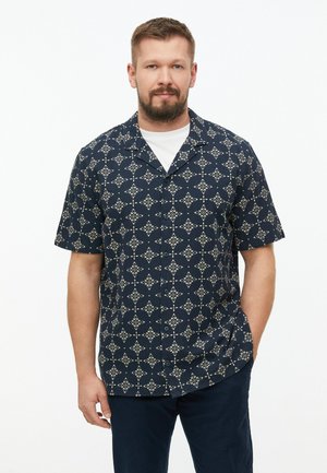 Homme barbu portant une chemise bleu marine à manches courtes avec un motif géométrique blanc et un sous-vêtement blanc, debout devant un fond blanc.