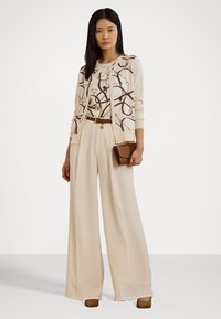 Cardigan bej tricotat cu accente aurii, purtat peste o bluză cu model, asortat cu pantaloni largi crema și o poșetă maro. Tocuri înalte maro.