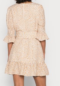 Donna che indossa un mini abito beige con stampa floreale bianca e orlo arricciato, vista da dietro su uno sfondo neutro.