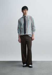 Άνδρας με τσαλακωμένο ριγέ πουκάμισο και μαύρη γραβάτα, καφέ παντελόνι φαρδιάς γραμμής και ασπρόμαυρα παπούτσια, στέκεται μπροστά σε ένα απλό φόντο.