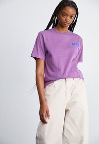 T-shirt en coton violet à manches courtes et à coupe décontractée, avec un logo bleu sur la poitrine gauche. Associé à un pantalon beige à jambes larges.