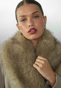 Beige kabát nagy, puha, világosbarna műszőrme gallérral. A modell az egyik kezével fogja a gallért, sima bőrrel és természetes színű ajakkal.