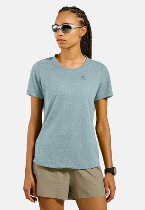 Femme portant un T-shirt bleu clair à manches courtes, un short beige, des lunettes de soleil et une montre noire au poignet, debout devant un fond blanc.