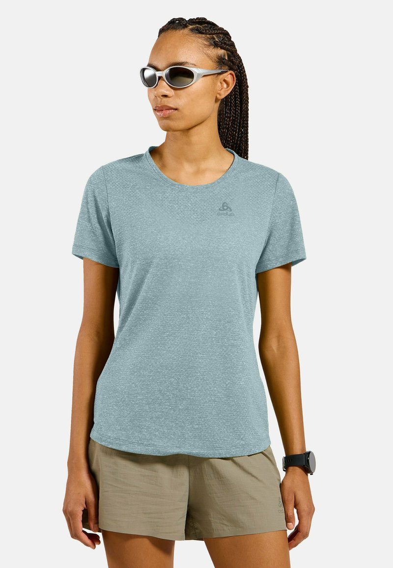 Frau trägt hellblaues T-Shirt mit kurzen Ärmeln, beige Shorts, Sonnenbrille und schwarze Armbanduhr, steht vor weißem Hintergrund.