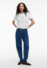 Witte katoenen t-shirt met een zwarte lijntekening van een gezicht en bloemen. Gepaard met donkerblauwe wijdvallende spijkerbroek en bruine schoenen.