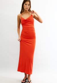 Robe maxi sans manches rouge avec un décolleté drapé, silhouette ajustée et texture lisse, atteignant la longueur des chevilles. Accentuée par des bretelles spaghetti.