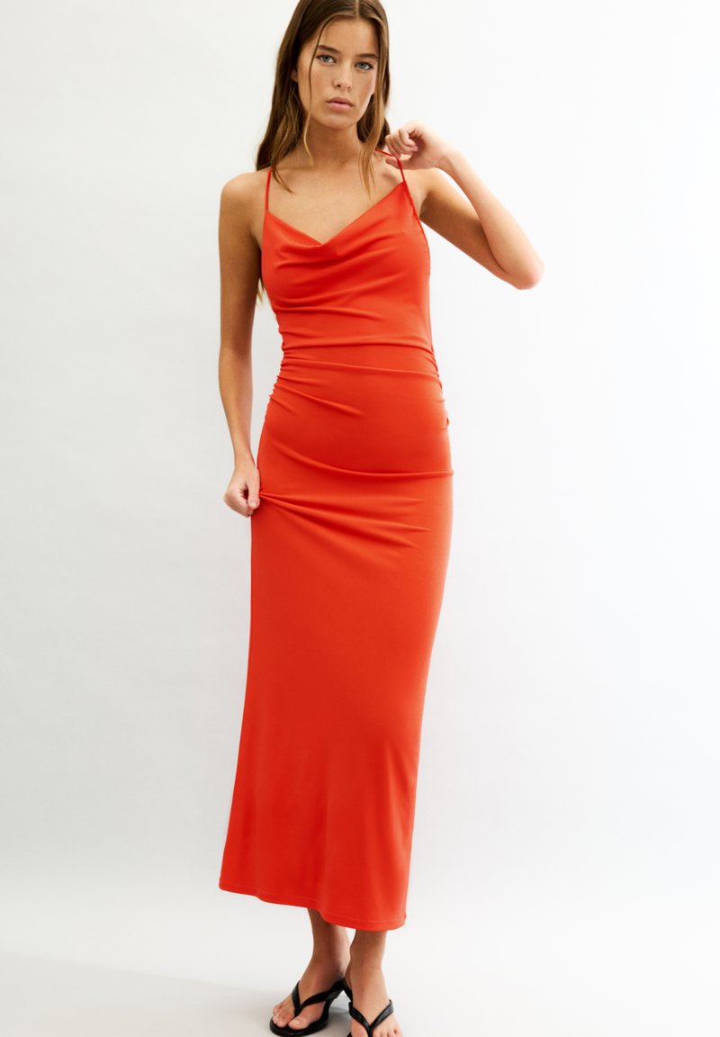Robe maxi sans manches rouge avec un décolleté drapé, silhouette ajustée et texture lisse, atteignant la longueur des chevilles. Accentuée par des bretelles spaghetti.