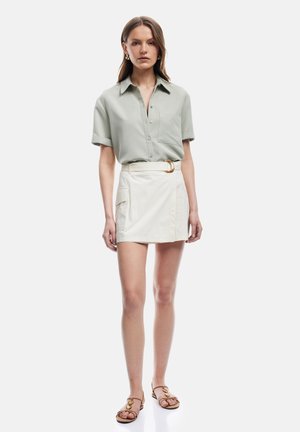 Vrouw draagt een lichtgrijze korte mouwen blouse met knopen, een witte minirok met riem en zak, en bruine platte sandalen, staand tegen een witte achtergrond.