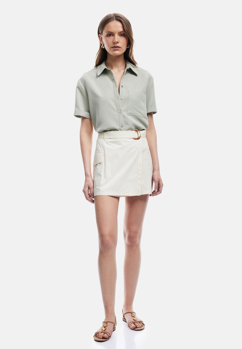 Mujer con camisa de manga corta de color gris claro con botones, minifalda blanca con cinturón y bolsillo, y sandalias planas marrones, de pie contra un fondo blanco.