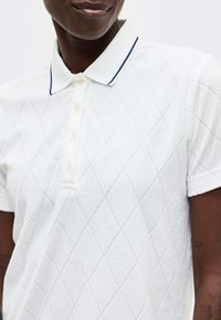 Άτομο που φοράει λευκό πόλο με κοντά μανίκια, διακριτικό διαμαντένιο μοτίβο και γιακά με μπλε περίγραμμα σε απλό φόντο.