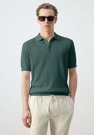 Hombre con camisa polo de manga corta de canalé verde oscuro, pantalones beige con cordón y gafas de sol negras, de pie frente a un fondo liso.