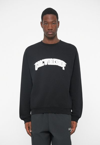 Ung mand iført en sort sweatshirt med hvid tekst "OSRC WORKSHOP", stående foran en ensfarvet hvid baggrund, foranvendt.