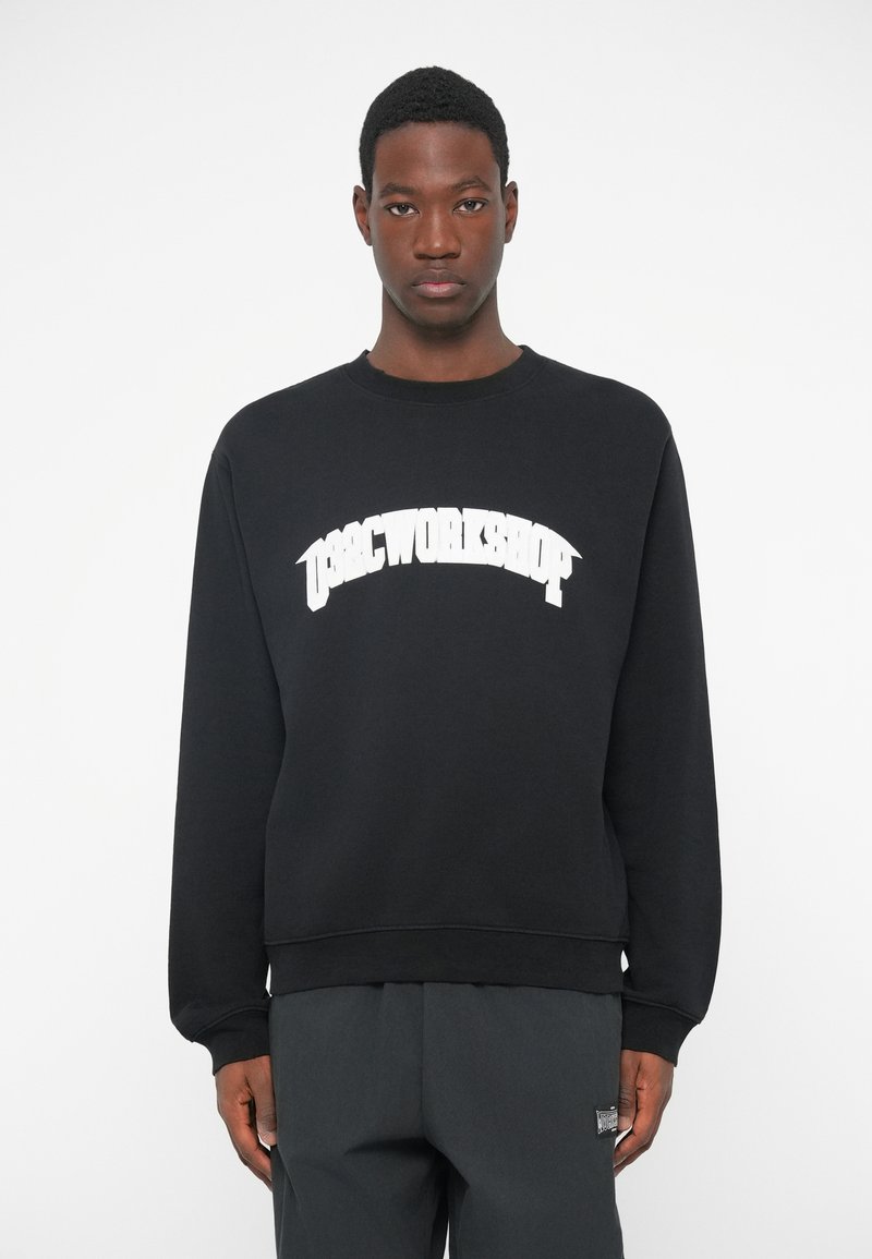 Jongeman in een zwarte sweatshirt met witte tekst "OSRC WORKSHOP", staande tegen een effen witte achtergrond, recht vooruit kijkend.