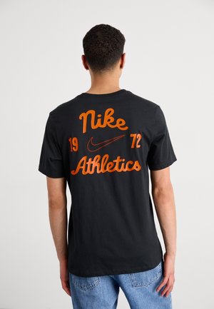 Osoba ubrana w czarną koszulkę Nike Athletics z pomarańczowym napisem i logo, w połączeniu z jasnoniebieskimi dżinsami, stojąca tyłem do kamery.