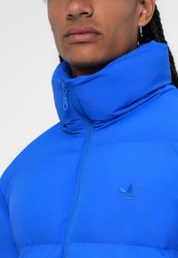 Modra puffer jakna z visokim ovratnikom, polnim zadrgo in diskretnim logotipom na prsih. Material se zdi gladek z lahko teksturo.