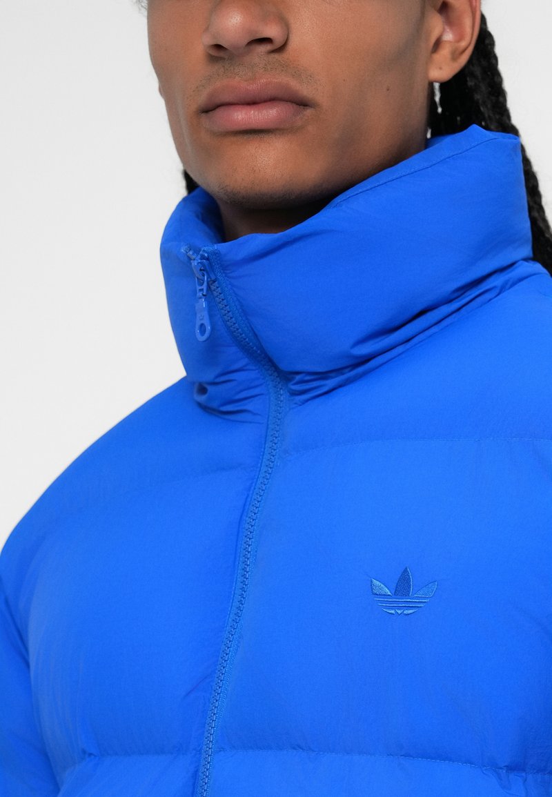 Giacca imbottita blu con alto colletto, zip completa e logo discreto sul petto. Il materiale sembra liscio con una texture leggera.