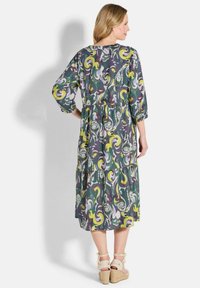 Frau, die ein Midikleid mit grünem, gelbem und lila abstraktem Muster, Dreiviertelärmel und beigen Keilabsätzen mit Knöchelbindungen trägt.