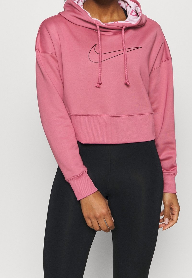 Personne portant un sweat à capuche court rose avec le logo Nike swoosh noir et des leggings noirs ajustés, debout devant un fond clair uni.