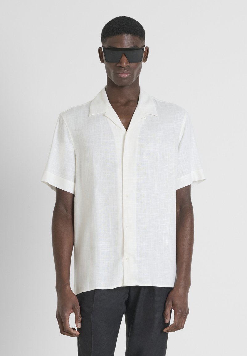 Camisa blanca de manga corta con botones y tejido texturizado, que presenta un cuello con muesca y un sutil detalle de paneles. Lleva puestas gafas de sol negras.