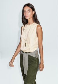 Top senza maniche color crema abbinato a pantaloni verde oliva. Un maglione grigio chiaro legato in vita. Design semplice, minimi accenti, texture liscia.