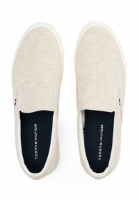 Beżowe buty wsuwane z teksturowanej tkaniny, biała gumowa podeszwa i granatowe wnętrze z nazwą marki "TOMMY HILFIGER."