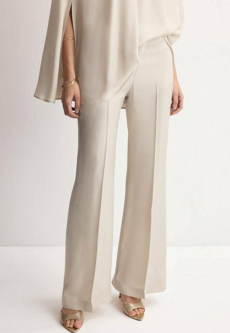 Femme portant un pantalon large beige, un haut ample assorti, des bracelets en or et des talons ouverts dorés, debout devant un fond uni.