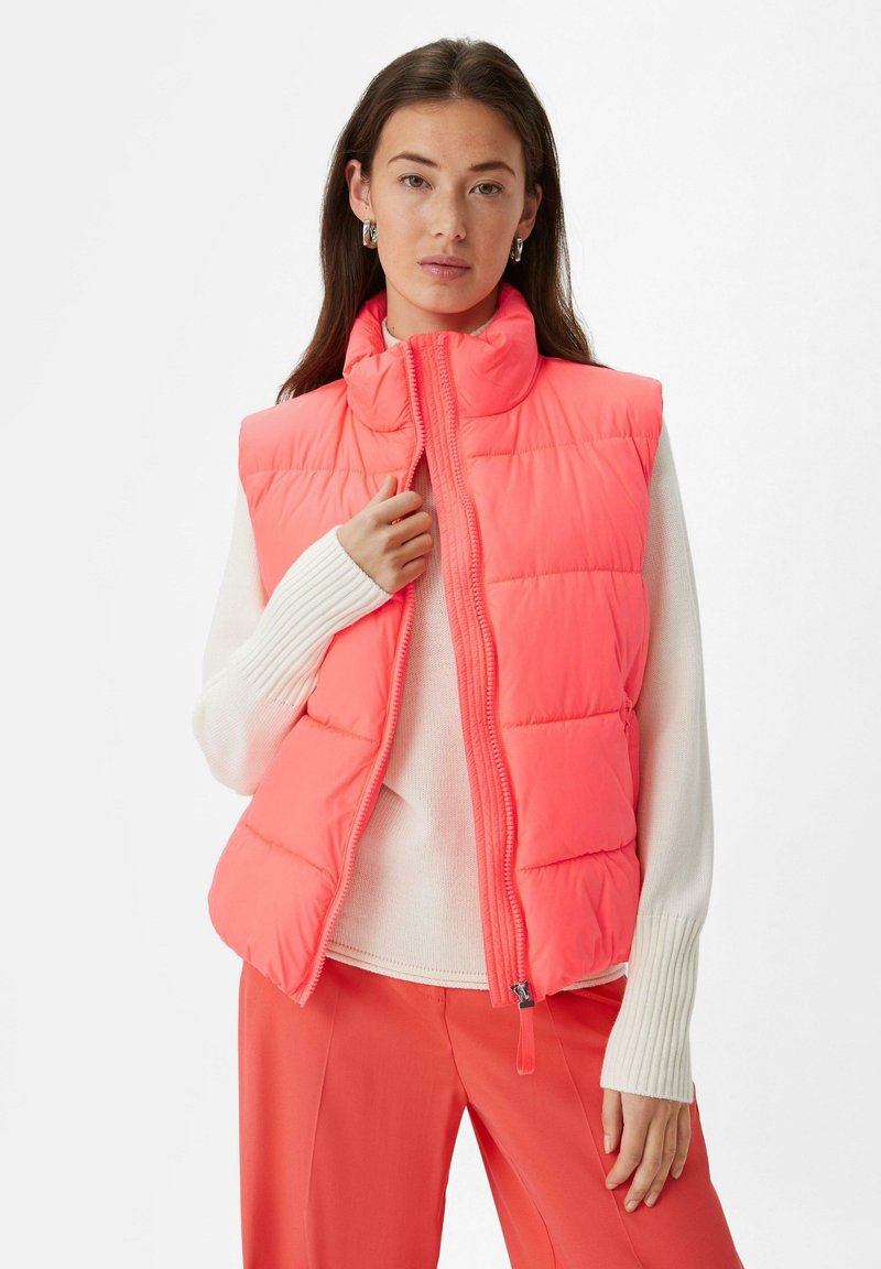 Gepolsterte ärmellose Weste in leuchtendem Korallenrot, mit hohem Kragen, vorderem Reißverschluss und gestepptem Design, drüber getragen über einem weißen Pullover.