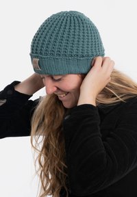 Spleciona w kolorze turkusowym czapka z ribbowym mankietem i tkanym wzorem, z małą metką z tkaniny. Modelka ma długie włosy i nosi czarną kurtkę ze sztruksu.