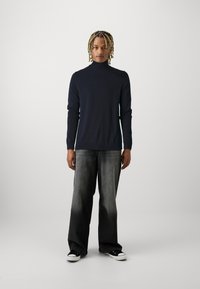 Suéter de gola alta azul-marinho com mangas longas, combinado com umas calças largas em degradê preto e cinza e sapatilhas pretas com solas brancas.