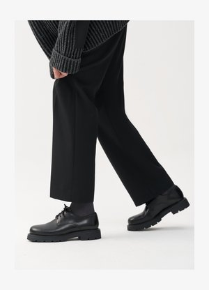 Black leather shoes with a rounded toe, lace-up design, and thick, textured sole. Worn with tailored black trousers and a ribbed sweater.