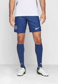 Blauwe sportshorts met verticale strepen, voorzien van een Parijs-logo. Gecombineerd met blauwe sokken en witte voetbalschoenen met Nike-merk. 